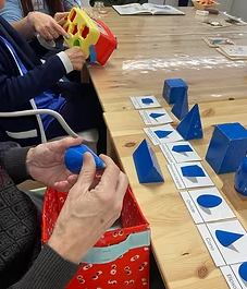 Personas mayores participando en actividades de estimulación cognitiva con materiales Montessori. Se observa a una persona sosteniendo una figura geométrica azul y explorándola con las manos, mientras otra inserta formas en un juego de encaje. En la mesa hay tarjetas con nombres de figuras geométricas y distintos materiales diseñados para ejercitar la memoria y la motricidad fina